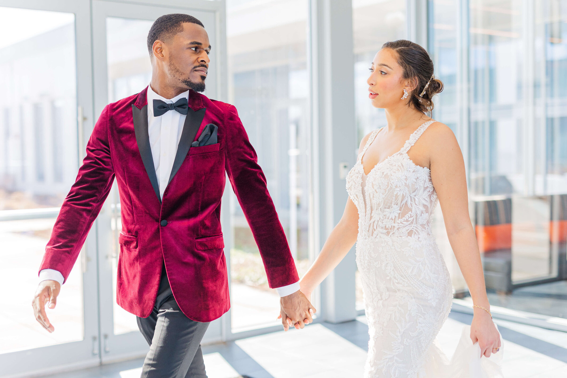 A groom in a maroon jacket holds his bride's hand and guides her as they walk through a hotel with a lot of windows. they are gazing at each other.