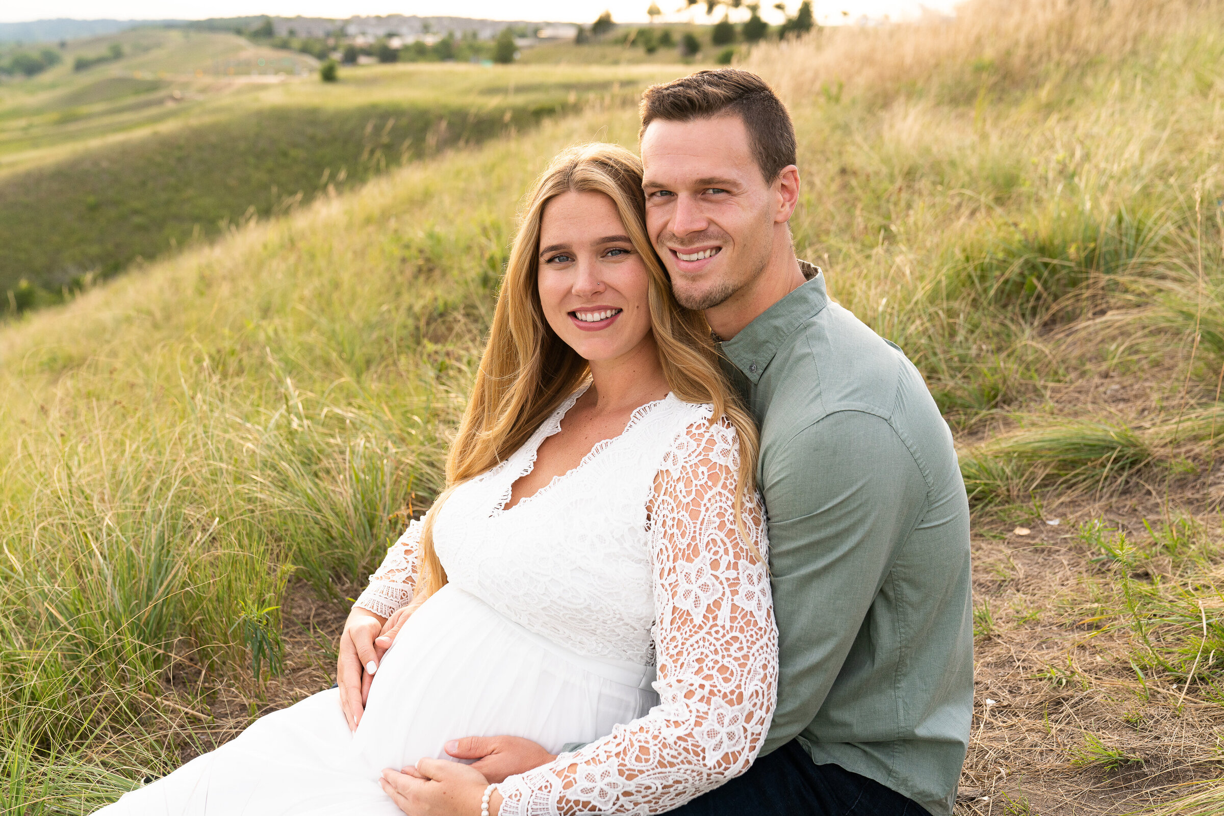 Couple sit in the tall grass and hold the woman's baby belly