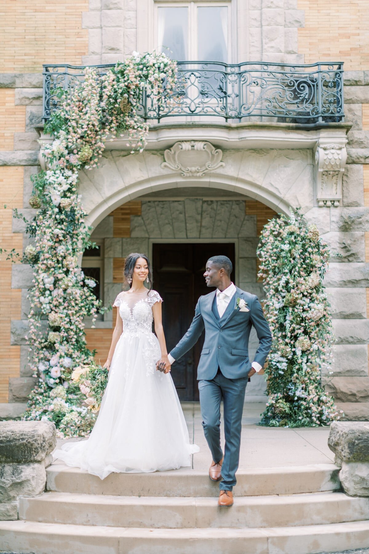 Bride and groom walk hand in hand down stairs of a stunning, brick venue with a flower arch.
