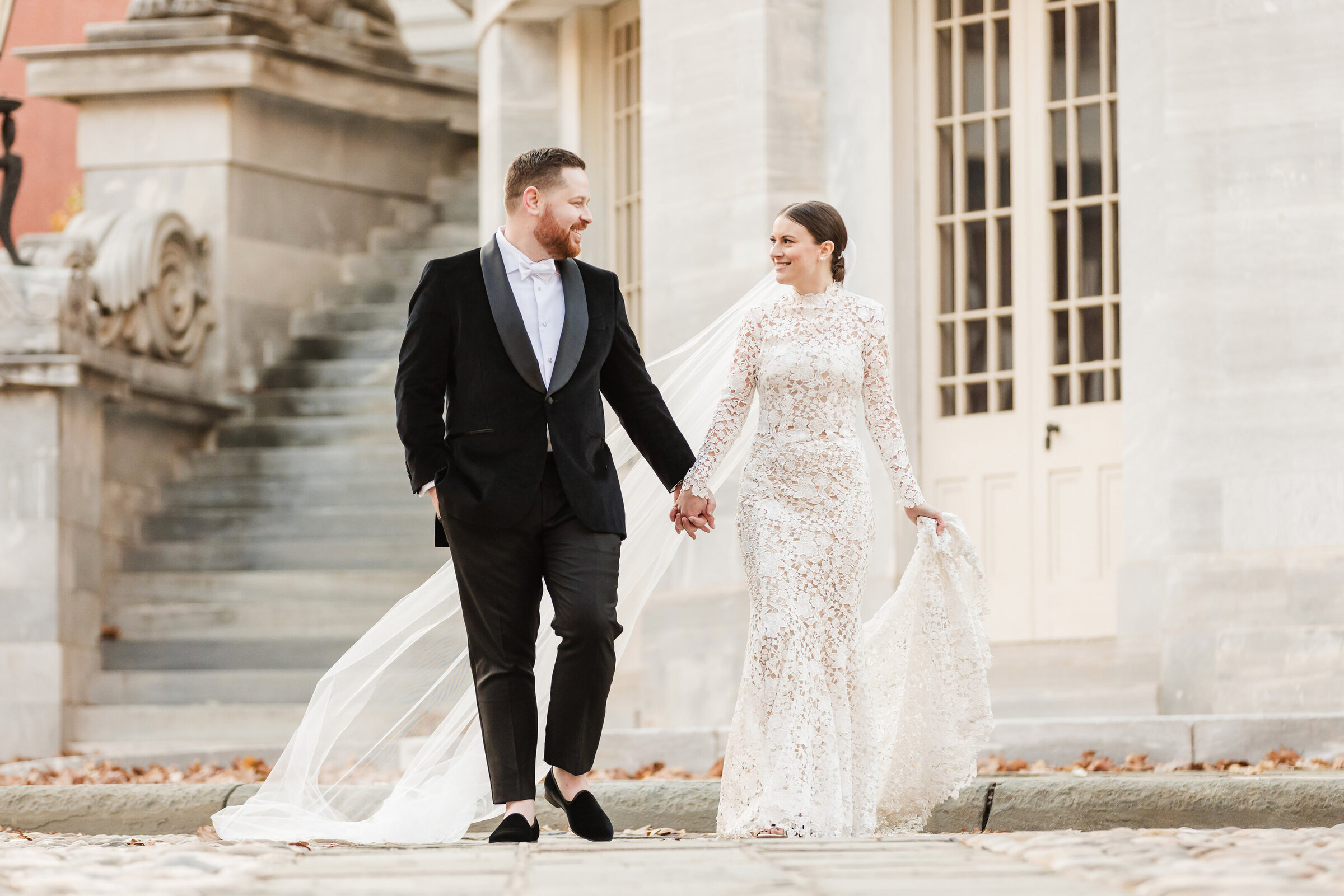 Bride wearing a long sleeve Monique Lhuillier  lace gown is walking with her groom holding hands at Merchants Exchange Building on their wedding in Philadelphia.