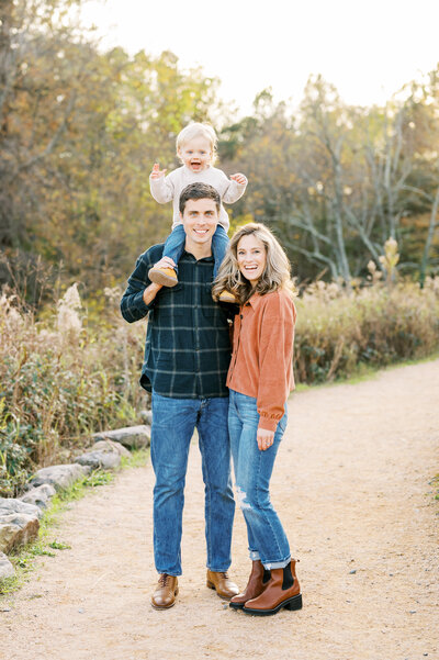 Family smiles at camera with little boy on dad's shoulders during outdoor photo session in Raleigh NC