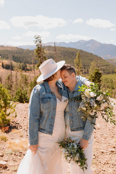 Bride with white hat kisses her bride's forehead while both hold bouquets with Rocky Mountains behind them.