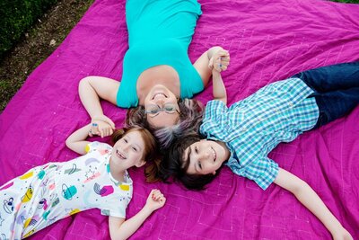 a mom and 2 kids lay on a pink blanket holding hands