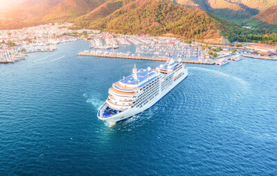 A large cruise ship sails through a bright blue sea, nearing a harbor lined with numerous boats. The coastal town is nestled against lush green hills, bathed in sunlight, under a clear sky.