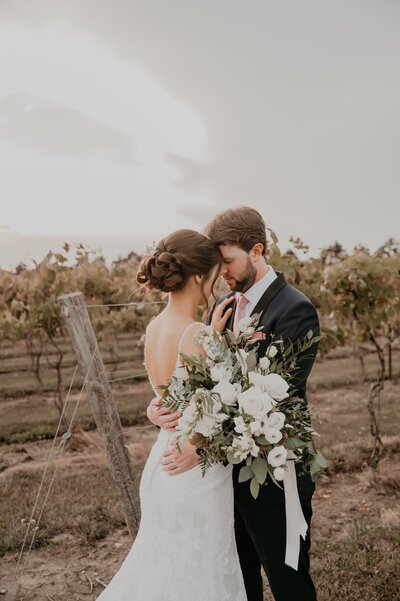 Bride and groom are wrapped in an embrace with their heads touching for a wedding day portrait photo. Behind them are grape vines at a Niagara, ON winery. Captured by top Niagara wedding photographer Ashlee Ellison.