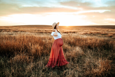 Pregnant woman closes eyes and tips head back at sunset in pink flowy skirt and white crop top with wide brim felt hat