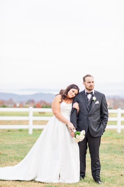 Bride wearing a Maggie Sottero gown and groom wearing a black tuxedo posing together sweetly in a fall field on their wedding day.