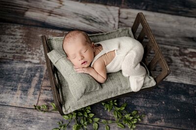 newborn baby boy dressed in cream knit overalls is posed on a bed with green  and brown accents
