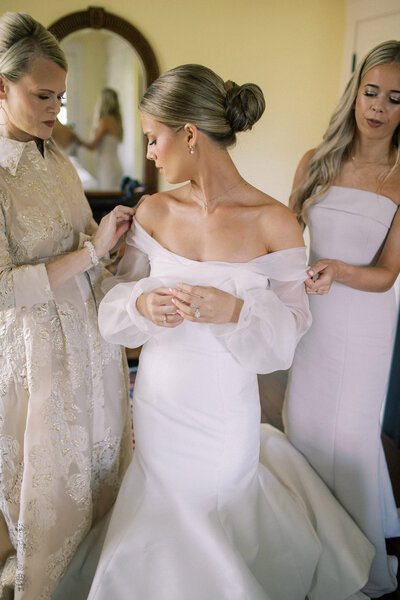 Sophisticated bride puts on her wedding gown with her mother and sister assisting