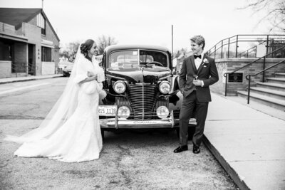 Stylish bride and groom pose with their vintage car in front of a church.