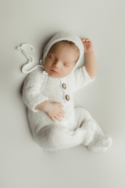 A newborn boy sleeping on his back wearing a white knitted romper in a beautiful studio setting