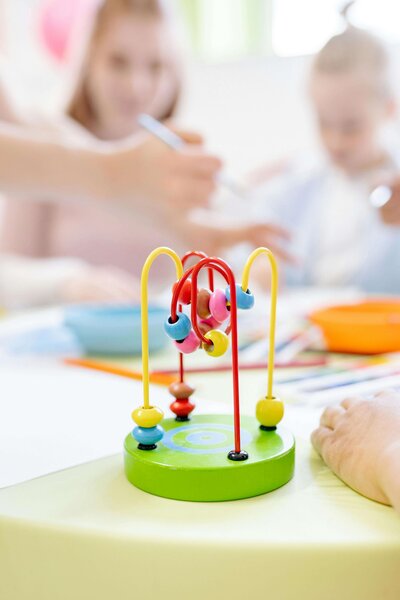 A colorful bead maze toy is placed on a table in the foreground, while blurred figures of children engaging in activities are visible in the background.