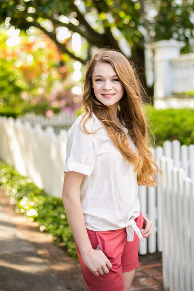 Teen girl in white blouse and pink shorts smiles in front of white picket fence