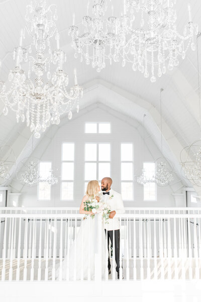 Bride and groom kiss on balcony of white barn wedding venue with crystal chandeliers above