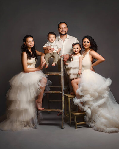 Fine art family portrait on gray background wearing gowns and dress attire in white, mom dad and three children two girls and a baby boy, posed with vintage ladders in my Phoenix Family studio