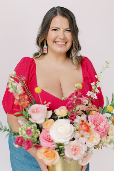 Brunette woman wearing hot pink shirt, holding brightly colored floral arrangement smiling at the camera