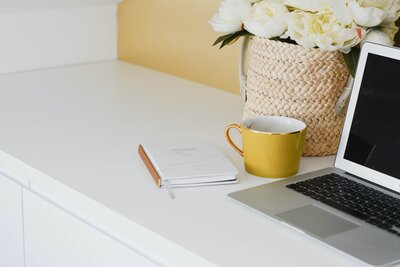 Laptop on a table with a yellow mug and a basket of white flowers