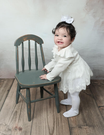 A joyful one-year-old child stands beside a grey chair during a milestone photography session in a studio setting in Rochester, NY. The child has a bright smile, capturing the essence of their first year of life.