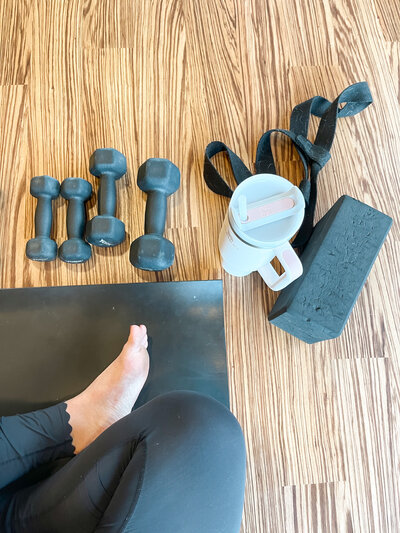 Woman's legs sitting on a yoga mat next to small dumbbells, a yoga block, and a Stanley water bottle