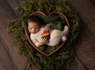 baby laying in a wooden heart bowl holding a rainbow heart
