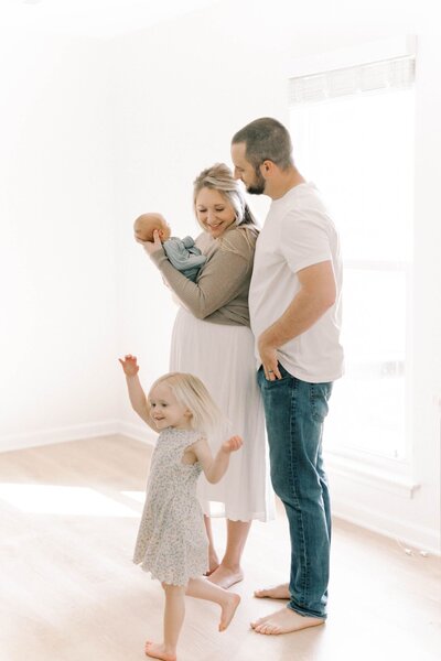 Mother holds newborn baby with dad  and daughter standing by playing.