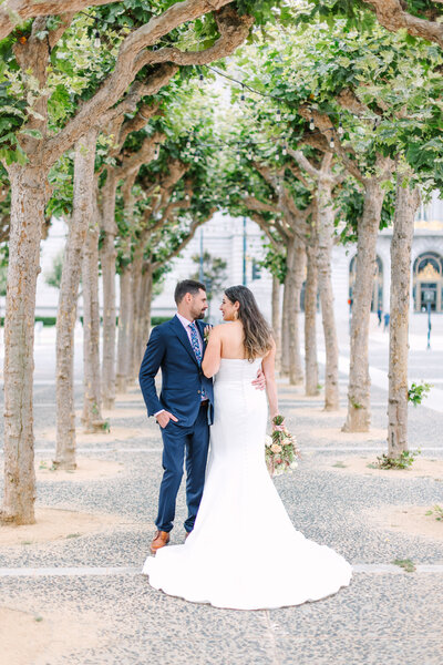 Bride and groom posing in an alley of trees outside of San Francisco City Hall wedding venue, bride wearing modern gown holding Jacqueline Ching Design bouquet, groom in navy suit with brown dress shoes, photo by Anastasiya Photography - San Francisco Wedding Photographer