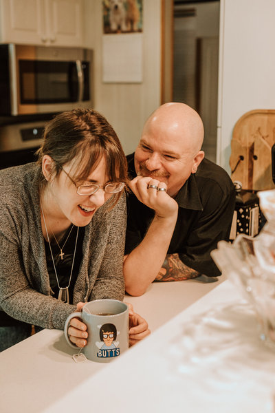 Man and woman laugh in a kitchen holding coffee cups.