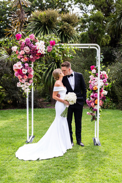 Married couple kissing under colourful flower arch