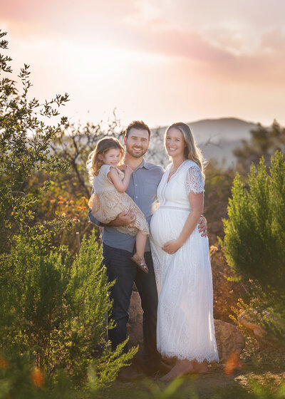 maternity photoshoot of mom wearing a white dress standing with dad and daughter smiling and holding baby bump in Los Angeles