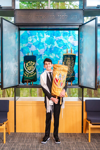 A teen boy in a black suit holds the large torah at the bimah