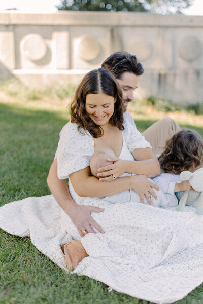 A mother, father, their toddler son and newborn baby sit on a white blanket outside during photo session with Boston newborn photographer Corinne Isabelle