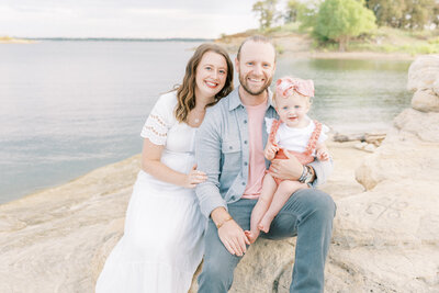 Family of 3 smiling at the camera by the water in murrell park with one year old baby girl photographed by Dallas Family Photographer Amanda Carter