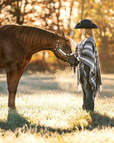 horse owner wearing cowboy hat and plaid poncho petting her chestnut american quarter horse mare in silver show halter in a field