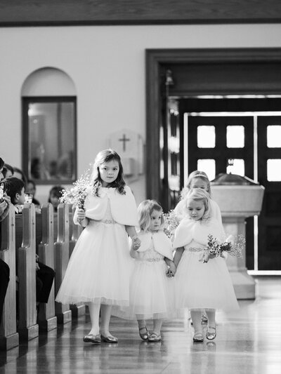 Four adorable flower girls dressed in matching white dresses hold hands as they nervously walk down the aisle in a catholic church