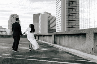 Newlyweds walk away from the camera holding hands on top of a parking garage in St. Louis, Missouri. Skyscrapers line the background.