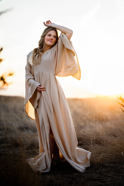 Senior girl wears long, flowy dress and puts her hand over her head at sunset