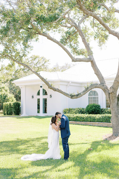 stunning white chapel  with bride and groom standing in the lawn in front of the building as the groom holds his brides hands and leans down to kiss them