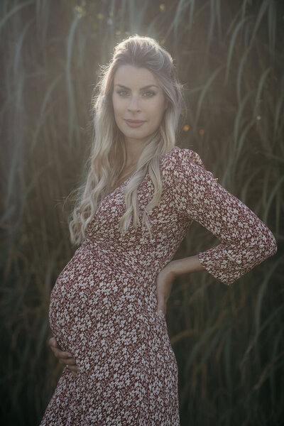 Eine schwangere Frau in einem blumigen Kleid steht vor einem Feld in der Sonne und präsentiert ihren Babybauch