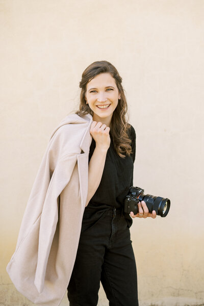 Winston-Salem wedding photographer, Jenn Eddine looks at the camera and smiles with a coat blazer over her shoulder and her camera in the other hand.