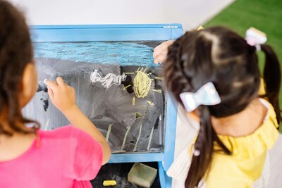 Two children are drawing with chalk on a small blackboard, creating colorful pictures that include a sun and clouds.