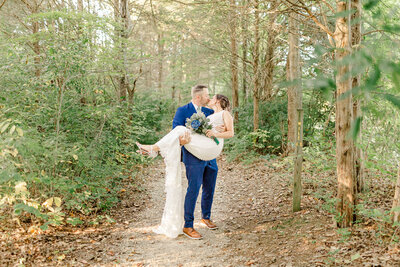 groom holding bride in his arms on a path in the woods while they kiss