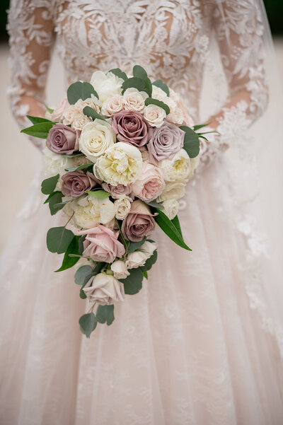A bride in an ornate lace dress holds a lush bridal bouquet of pale mauve, blush, and ivory roses accented with eucalyptus leaves