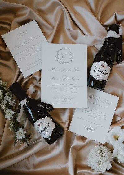 Wedding flat lay with elegant invitations, champagne mini bottles labeled Mr. and Mrs. Cook, and white florals on a satin fabric backdrop.