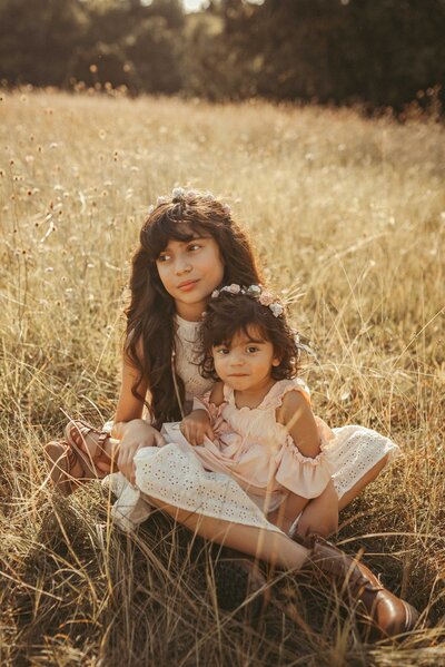 Two beautiful sisters captured in our Plano, Texas photography session, featured on our investment page. The sisters sit gracefully on the dried fall grass, wearing autumn dresses . The older sister cradles the youngest in her legs and have black hair.