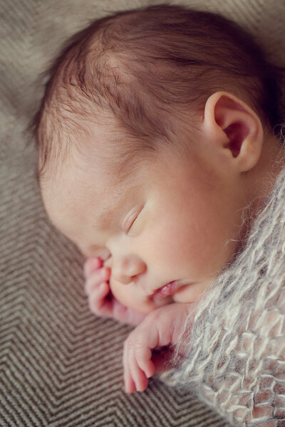 An adorable newborn is sleeping and wrapped in a super soft wrap with her hands peeking out the top.