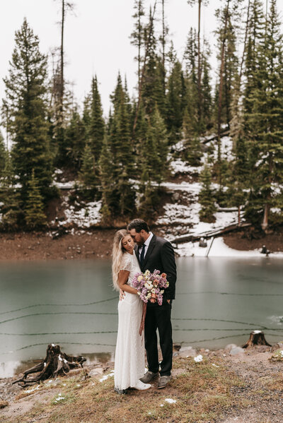 A couple stands close together near a frozen lake. A colorful floral bouquet in her hands.