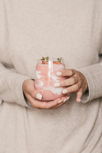 A woman holds a glass of strawberry yogurt, showcasing its pink color.