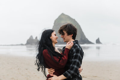 A couple stands face to face embracing in each other's arms in front of Haystack Rock at their Cannon Beach Engagement Photo Session. | Erica Swantek Photography