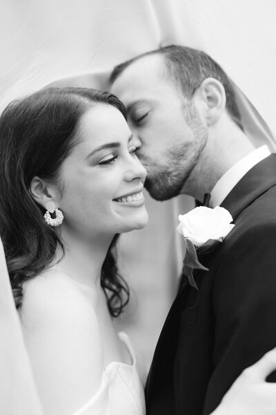 B&W timeless image of a newly married bride being kissed on the cheek by her groom.