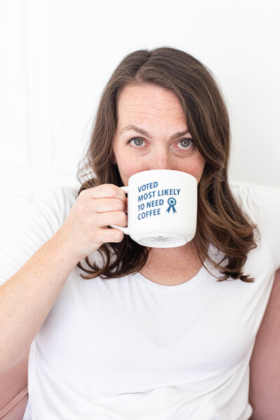 Photo of the social media strategist holding a white coffee mug with a dark blue inspirational quote in her right hand. she is covering half of her face with the mug